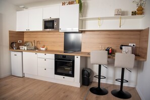 Eine kompakte Küche mit Mikrowelle, Backofen und Kaffeemaschine. Zwei Barhocker und ein kleiner Mülleimer sind vorhanden.