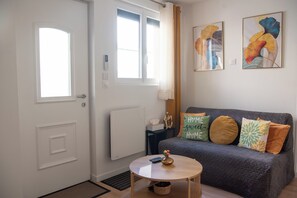 Una habitación con un sofá gris, una mesita redonda con un florero, una puerta con un ojo de cerradura y dos ventanas.
