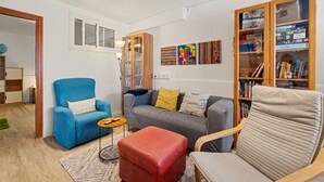 Ein Wohnzimmer mit blauem Sessel, grauem Sofa, Hocker in Rot und einem Holzregal voller Bücher und Puzzles.
