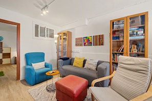 Ein Wohnzimmer mit blauem Sessel, grauem Sofa, Hocker in Rot und einem Holzregal voller Bücher und Puzzles.