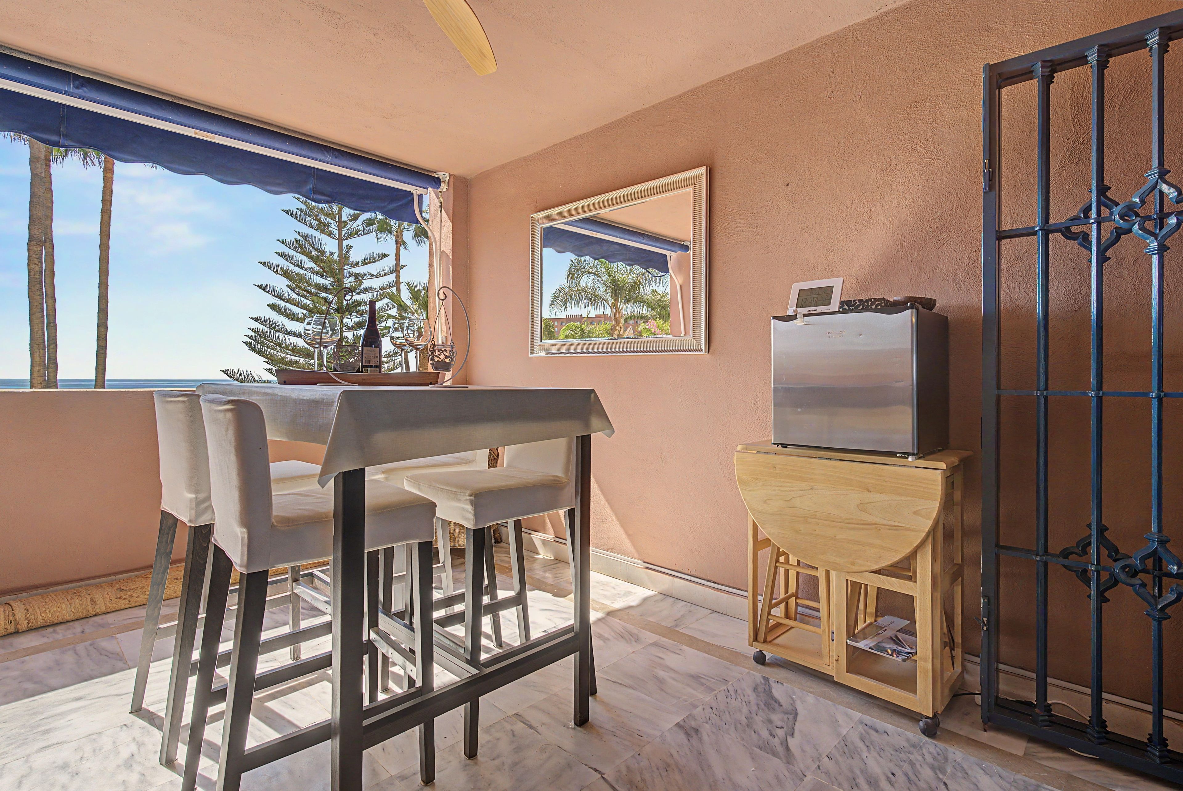 Una habitación con una mesa, sillas, un televisor sobre un soporte de madera, una ventana con vista a palmeras y una puerta con un portón metálico.