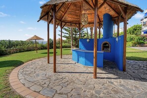 Un kiosque doté d’un toit de chaume, une structure bleue avec une cheminée, et une zone circulaire pavée de pierres.