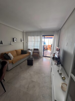 A modern living room with a sofa, armchair, and ottoman. There is a dining area with a table and chairs visible through a glass door.