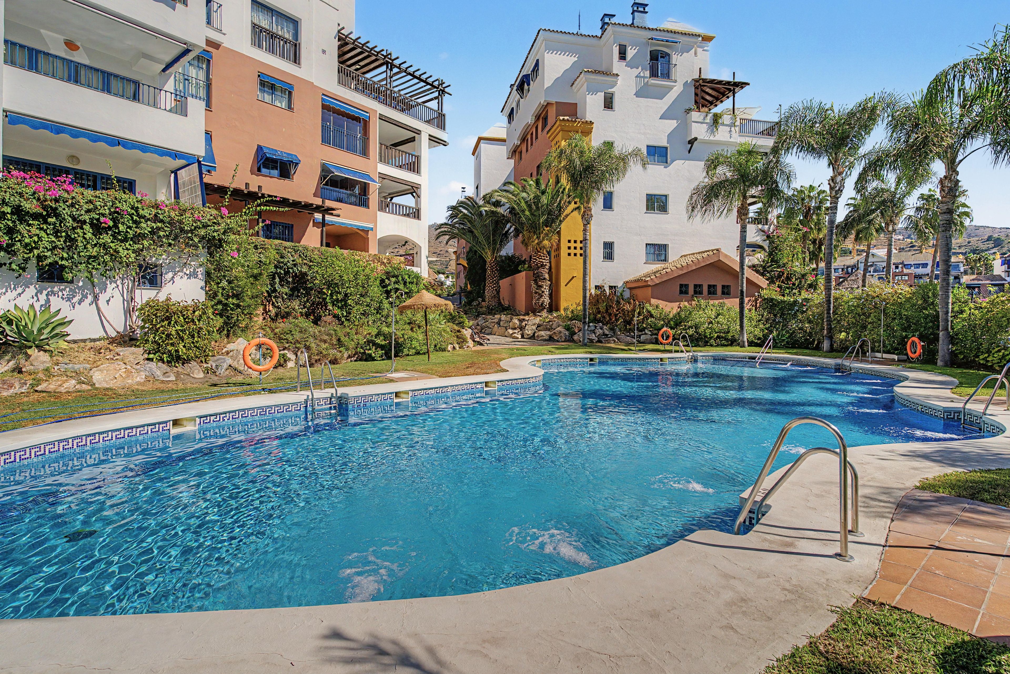 Ein Swimmingpool mit klarem, blauem Wasser, umgeben von einer Betonplattform, neben mehrstöckigen Gebäuden mit Balkonen.