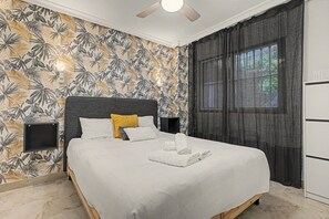 Habitación de hotel con cama, cabecera, mesita de noche, ventana con cortinas y pared con estampado de hojas tropicales.