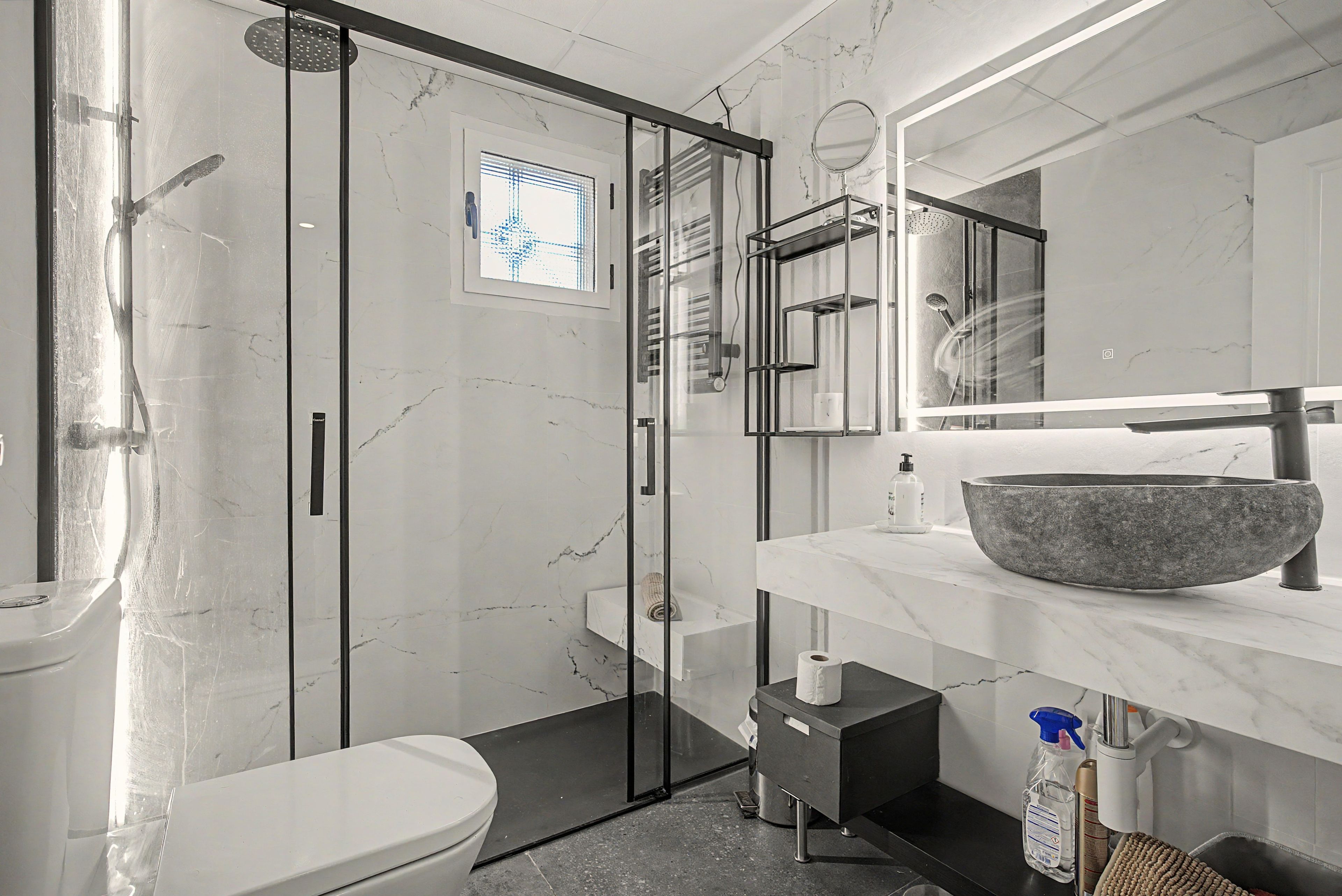 A modern bathroom with a glass-enclosed shower, a marble-tiled wall, and a large basin on the vanity.