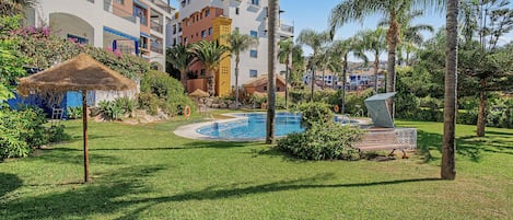 Un área de piscina con una sombrilla de techo de paja, palmeras y un edificio con balcones al fondo.