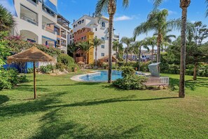 Un área de piscina con una sombrilla de techo de paja, palmeras y un edificio con balcones al fondo.
