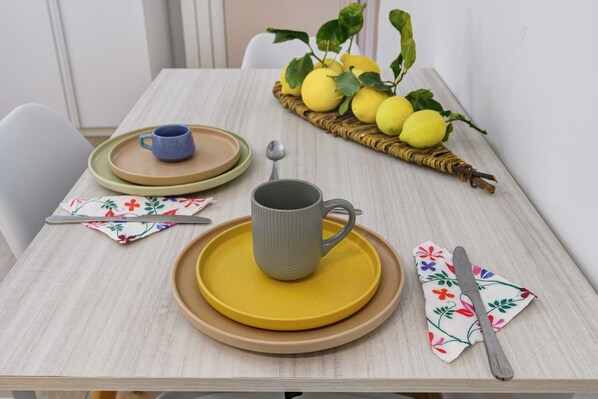 Uma mesa de jantar com um conjunto de pratos, uma caneca, uma colher, uma faca e um guardanapo estampado com um padrão floral. Há também uma cesta de vime com limões e uma planta.