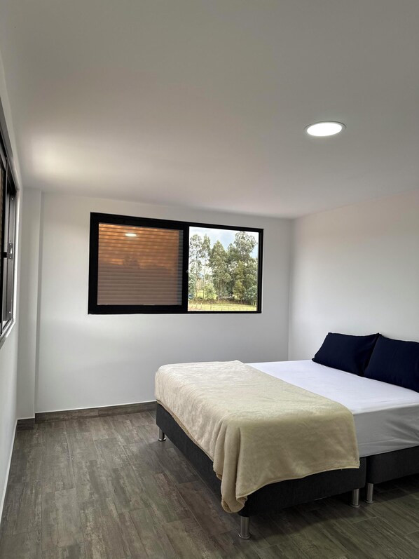 A bedroom with a bed, a window with blinds, a ceiling light, and a view of trees.