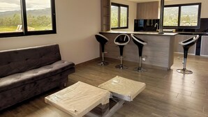 Una sala de estar moderna con un sofá, una zona de cocina con taburetes altos y grandes ventanales que ofrecen vistas al exterior.