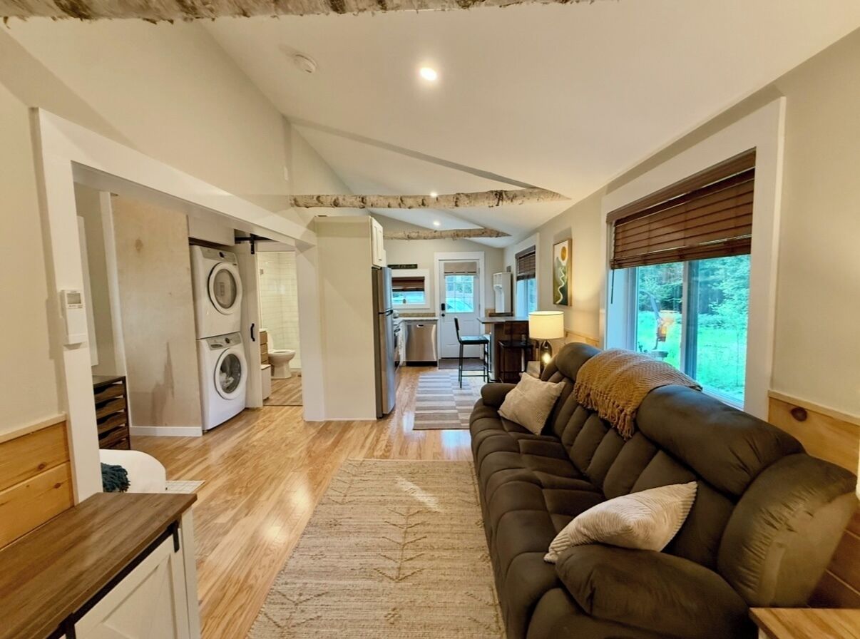 A living room with a sofa, a coffee table, and a rug. There is a washing machine and a kitchen in the background.