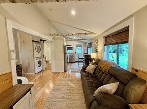 A living room with a sofa, a coffee table, and a rug. There is a washing machine and a kitchen in the background. - The Birch Retreat — Private Luxury Tiny Home in Bridgton, Maine with AC & WiFi (Bridgton)
