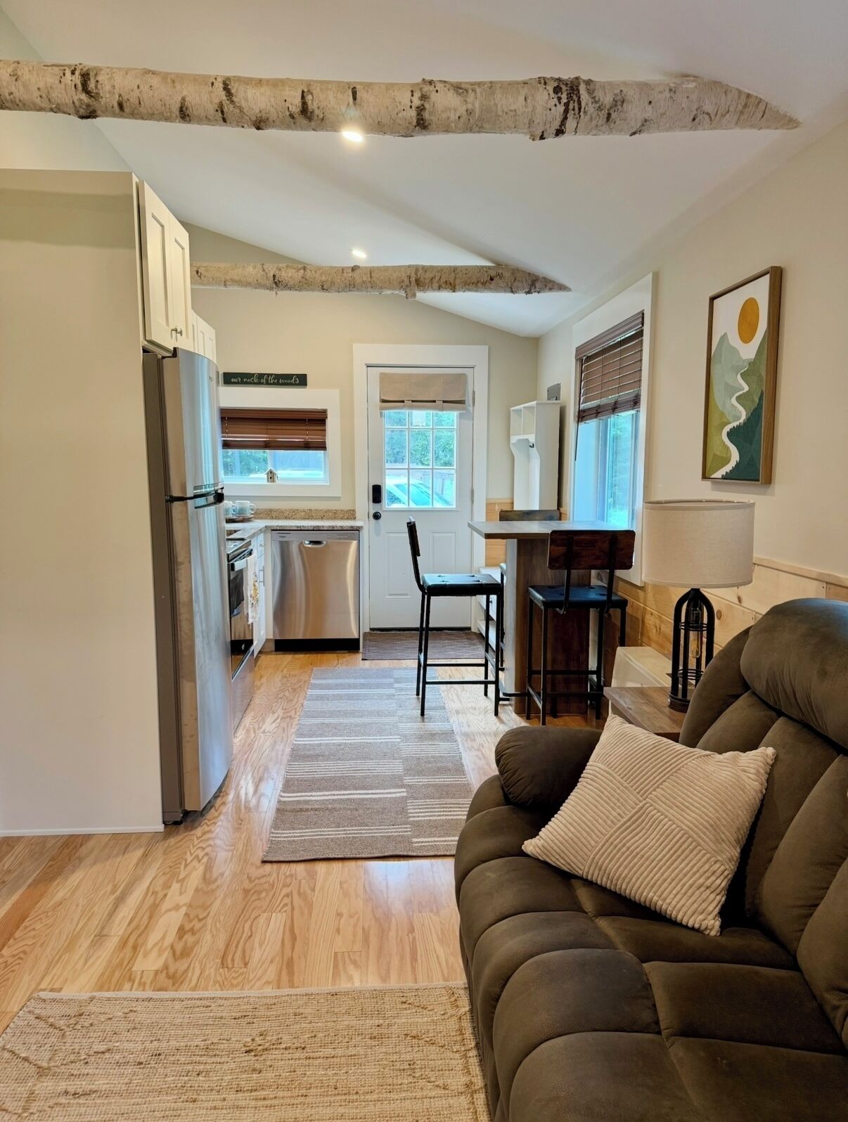 A modern kitchen with a refrigerator, dishwasher, and wooden cabinets. There is a dining area with a table and chairs, and a living room with a sofa and a rug.