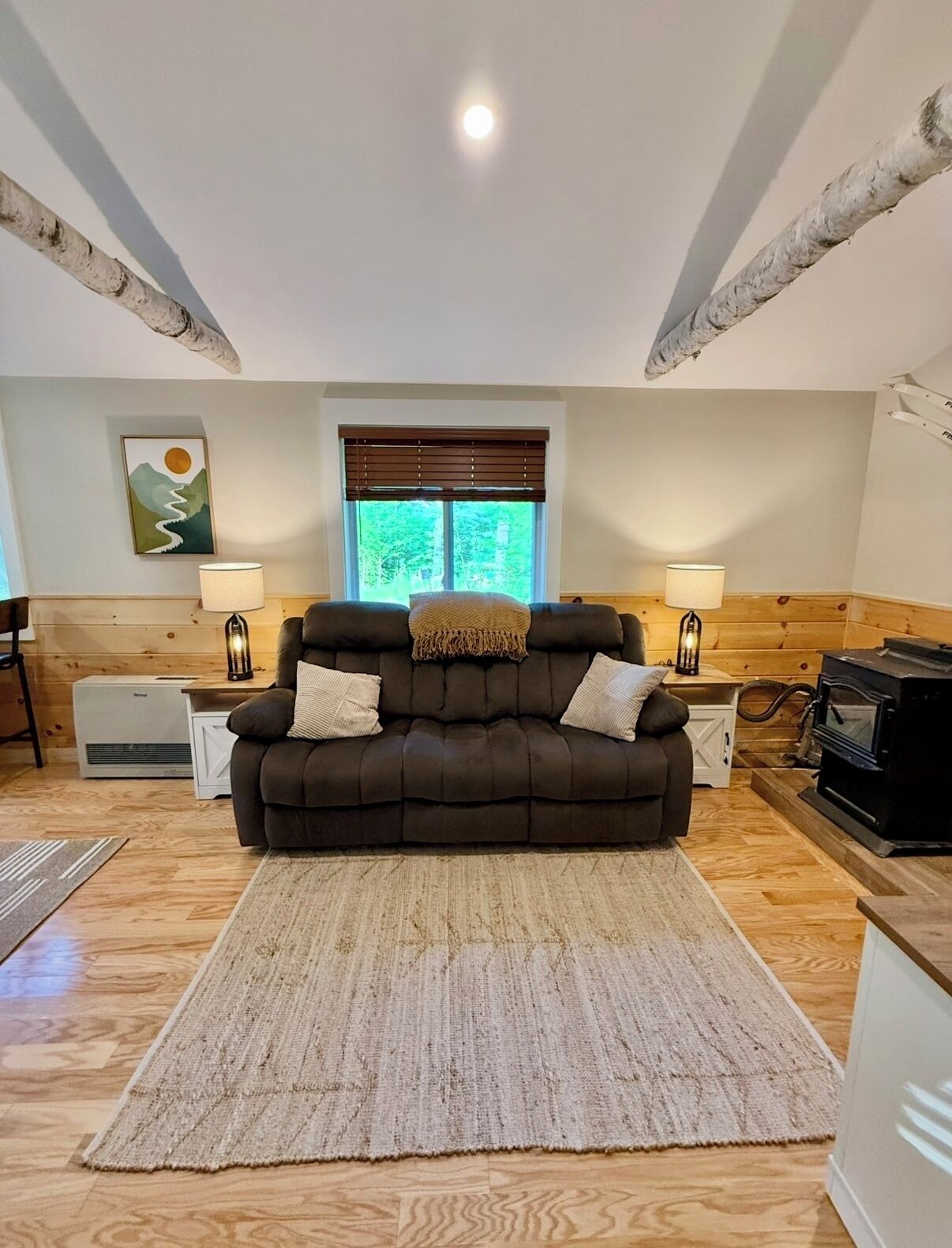 A cozy living room with a dark brown sofa, two side tables with lamps, a large window with blinds, a fireplace, and a wooden floor.