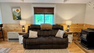 A cozy living room with a dark brown sofa, two side tables with lamps, a large window with blinds, a fireplace, and a wooden floor.