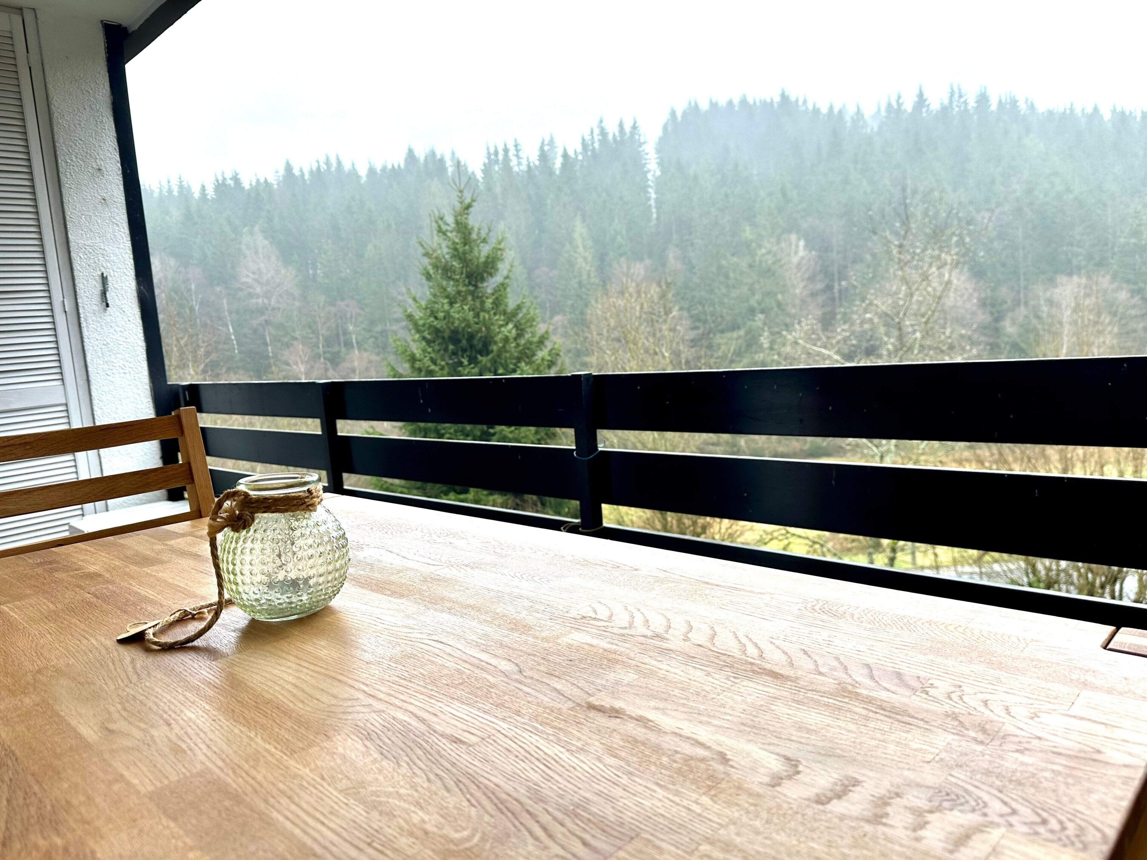 A wooden table with a glass jar and a pair of glasses on it, a black railing, and a view of a forested area.