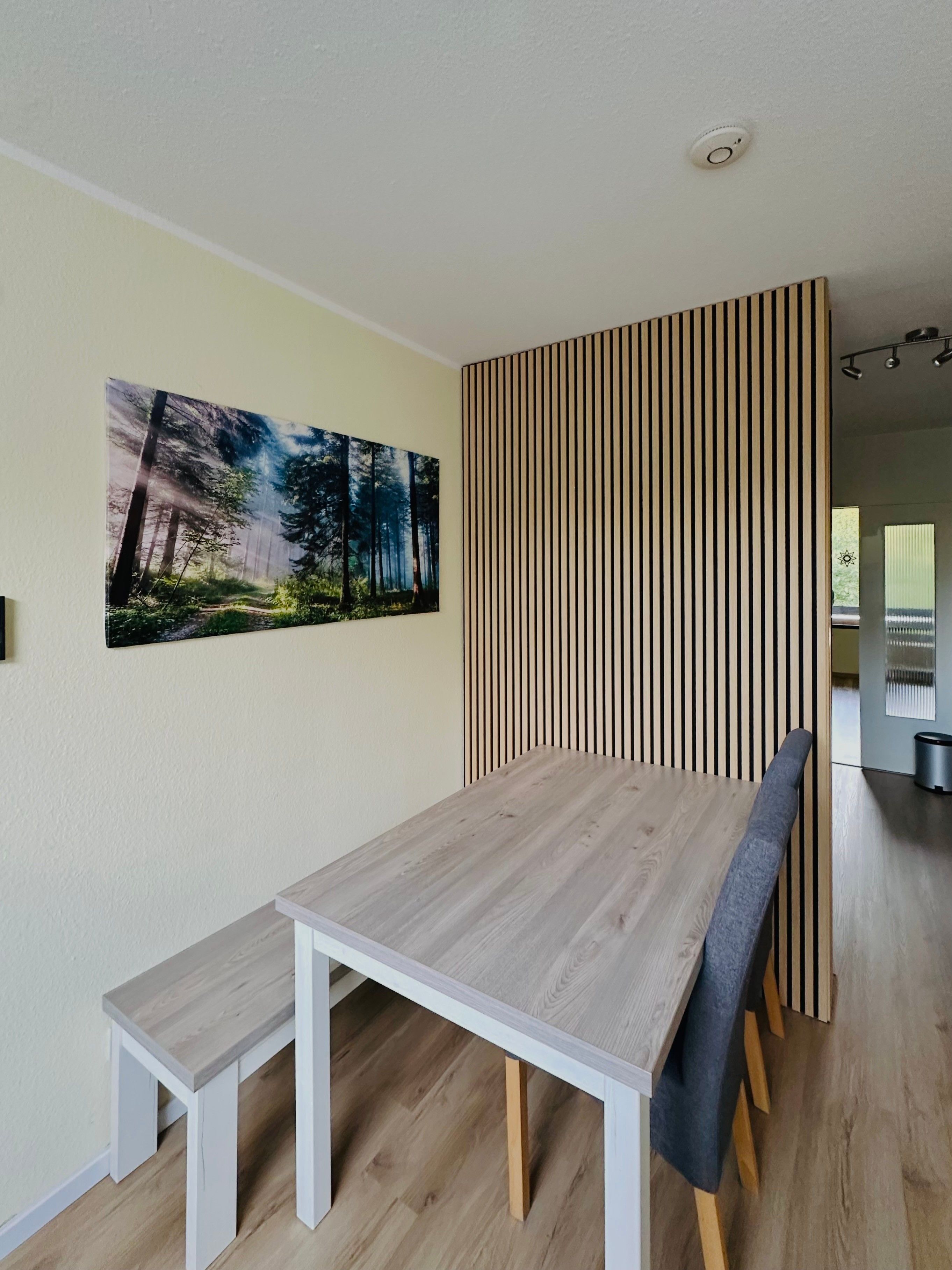 Un comedor con mesa y bancos de madera, un panel de madera vertical y un cuadro con motivos forestales.