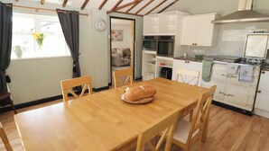 A dining area with a wooden table and chairs, a kitchen with white cabinets and appliances, and a window with curtains. - Farm Cottage (Carmarthen)
