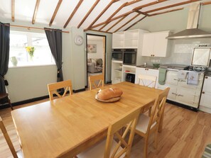 A dining area with a wooden table and chairs, a kitchen with white cabinets and appliances, and a window with curtains. - Farm Cottage (Carmarthen)