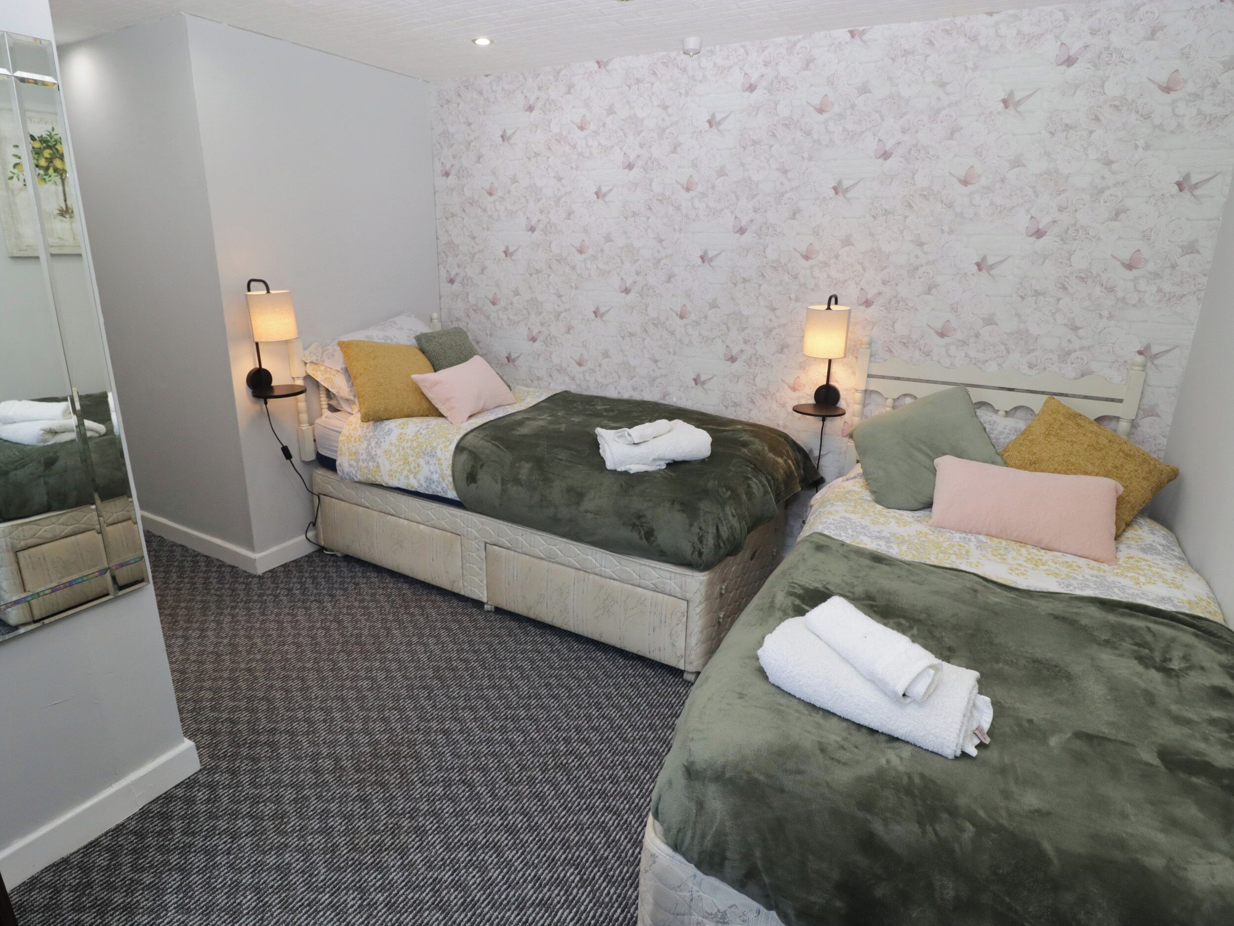 Ein Hotelzimmer mit zwei Betten, jeweils mit grüner Bettdecke und mehreren Kissen. An beiden Seiten der Betten befinden sich Wandlampen.