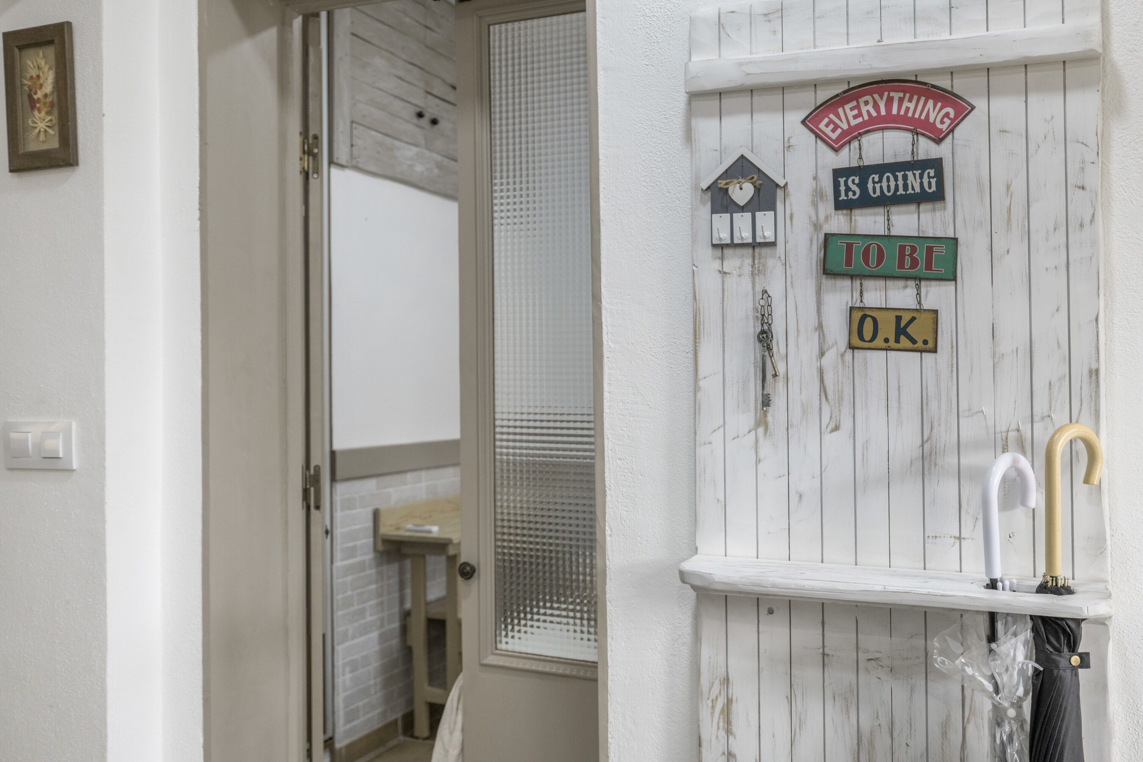Une porte blanche ornée d’un panneau en bois, une petite étagère avec un parapluie, et une pancarte sur laquelle on peut lire : « TOUT VA BIEN SE PASSER ».
