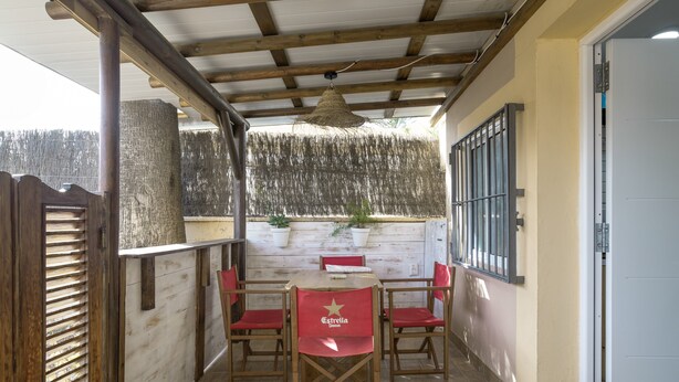 Un porche cubierto con una mesa de madera y sillas rojas, una lámpara de techo tejida y vistas a un área boscosa.