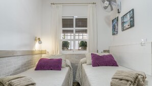 Una habitación con dos camas, una ventana con cortinas, un ventilador de pared y una lámpara fijada a la pared.