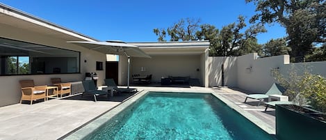 Un área de piscina moderna al aire libre con sillas de descanso, una barbacoa y un espacio de estar cubierto.