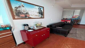 A living room with a red cabinet, a black sofa, a painting of a train, and a kitchen area with a refrigerator and a coffee maker. - MAISON COLLECTION PREMIUM (Porto Velho)
