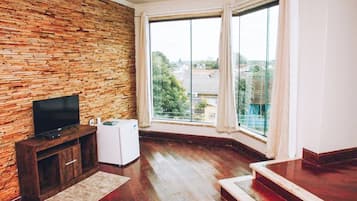 Quarto com parede de tijolos, televisão de tela plana, eletrodoméstico pequeno e uma janela grande com cortinas.
