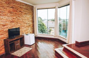 Una habitación con pared de ladrillo, un televisor de pantalla plana, un electrodoméstico blanco pequeño y un amplio ventanal con cortinas.