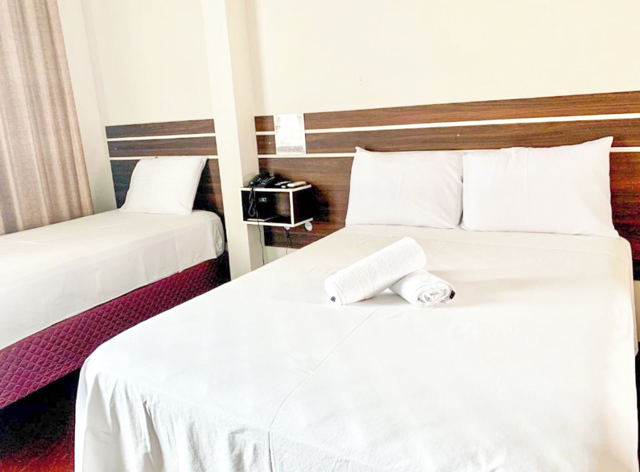 A hotel room with two beds, a wooden headboard, white linens, and a rolled towel on the larger bed.