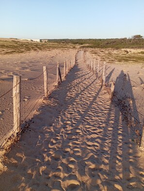 A sandy pathway bordered by wooden posts and barbed wire, leading towards a distant building.