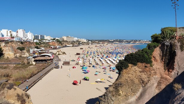 Een strand met rijen parasols en ligstoelen, een boulevard en gebouwen op de achtergrond.