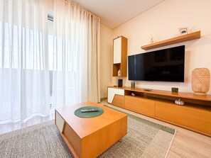 Ein modernes Wohnzimmer mit einem Holztisch, einem flachbildfernseher an der Wand und einem großen Fenster mit durchsichtigen Vorhängen.