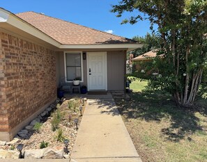 A residential house with a brick facade, a front door, a small porch with a chair and potted plants, and a pathway leading to the door. - Newly Updated 2BR/2BA • King Suite • Near ACU (Abilene)