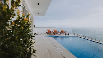 A swimming pool with lounge chairs, a clear view of the ocean, and a modern building with large windows.