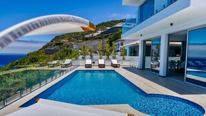 Un área de piscina moderna con sillas de descanso, vistas despejadas del océano y una colina al fondo.