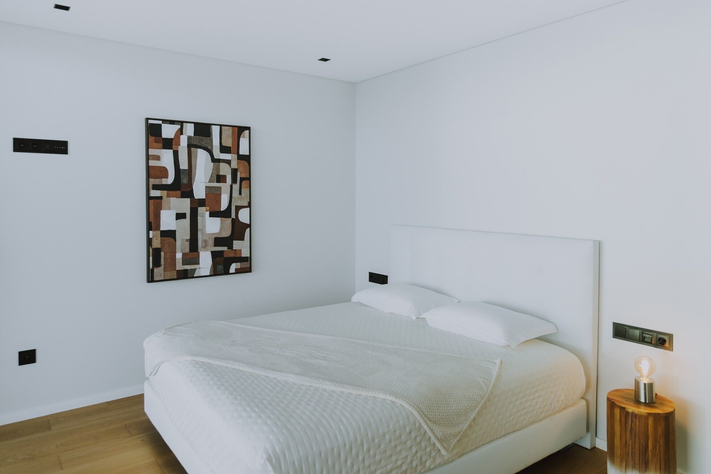 A modern bedroom with a large bed, a wall-mounted abstract art piece, and a bedside table with a lamp.