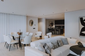A modern living room with a glass dining table, white chairs, a white sofa with patterned cushions, and a kitchen in the background. - Villa Nero – Private Luxusvilla mit beheizbarem Pool & Atlantikblick (Arco da Calheta)