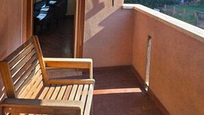 Un balcón con banca de madera, una puerta que da a un espacio interior y vistas a zonas verdes.