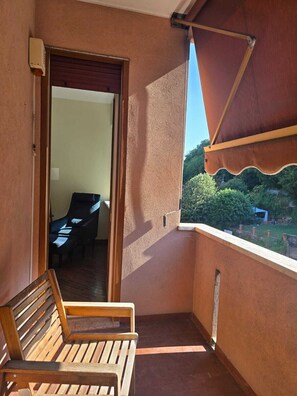 Ein Balkon mit einer hölzernen Sitzbank, einer Tür zu einem Innenbereich und Blick auf Grünfläche.