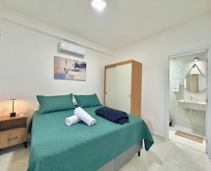 Ein Hotelzimmer mit Bett, Nachttisch mit Lampe, Wandklimaanlage, einem Bild, einem Spiegel und einem Badezimmer mit Waschbecken und Spiegel.
