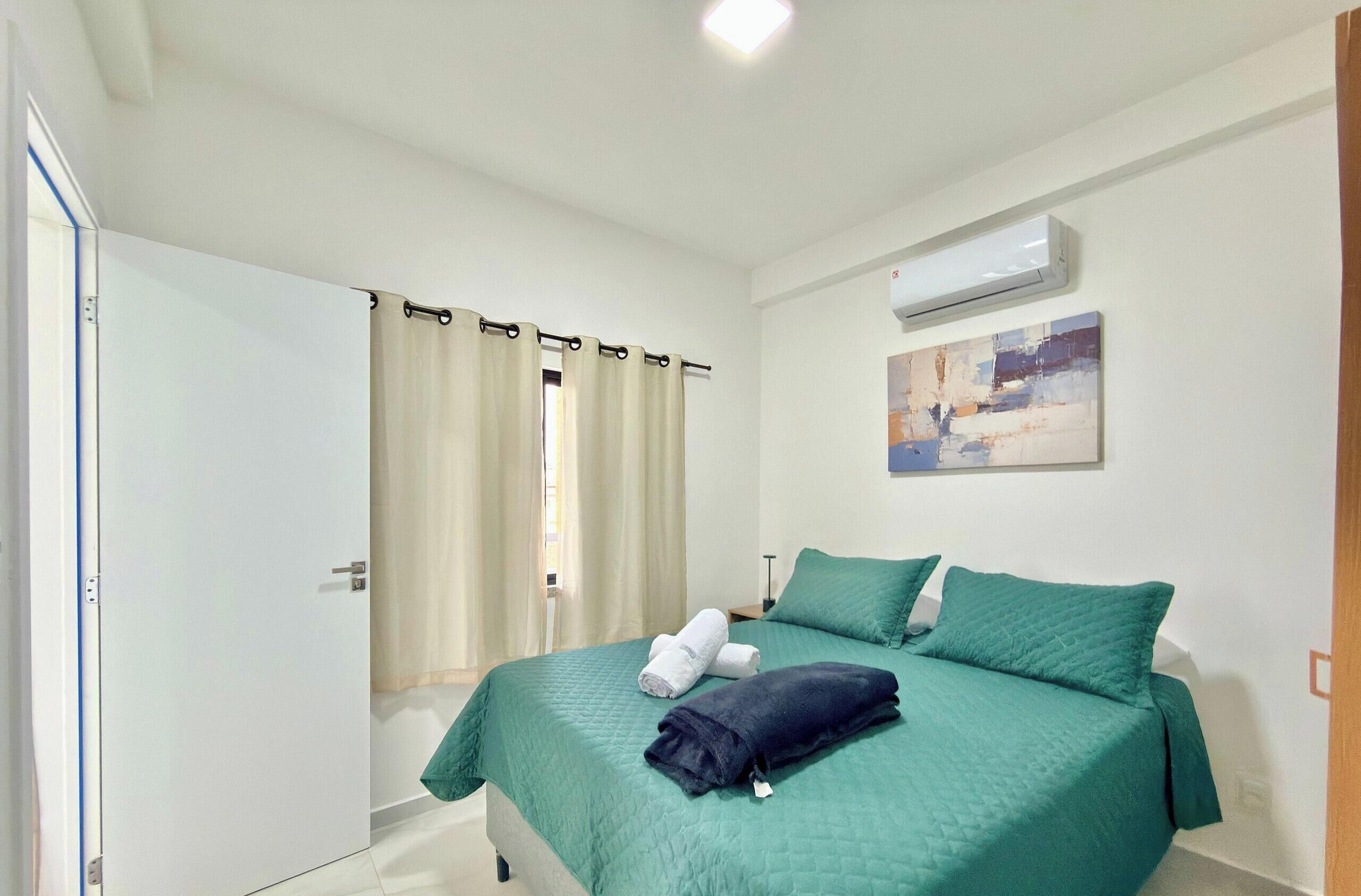 Una camera da letto con un letto, una finestra con tende, un condizionatore e un quadro appeso al muro.