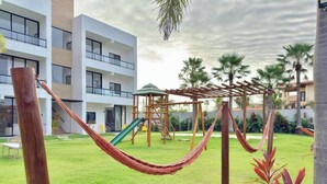 Een modern gebouw met een speeltuin, inclusief een glijbaan en klimconstructies, omringd door een gazon en palmbomen.
