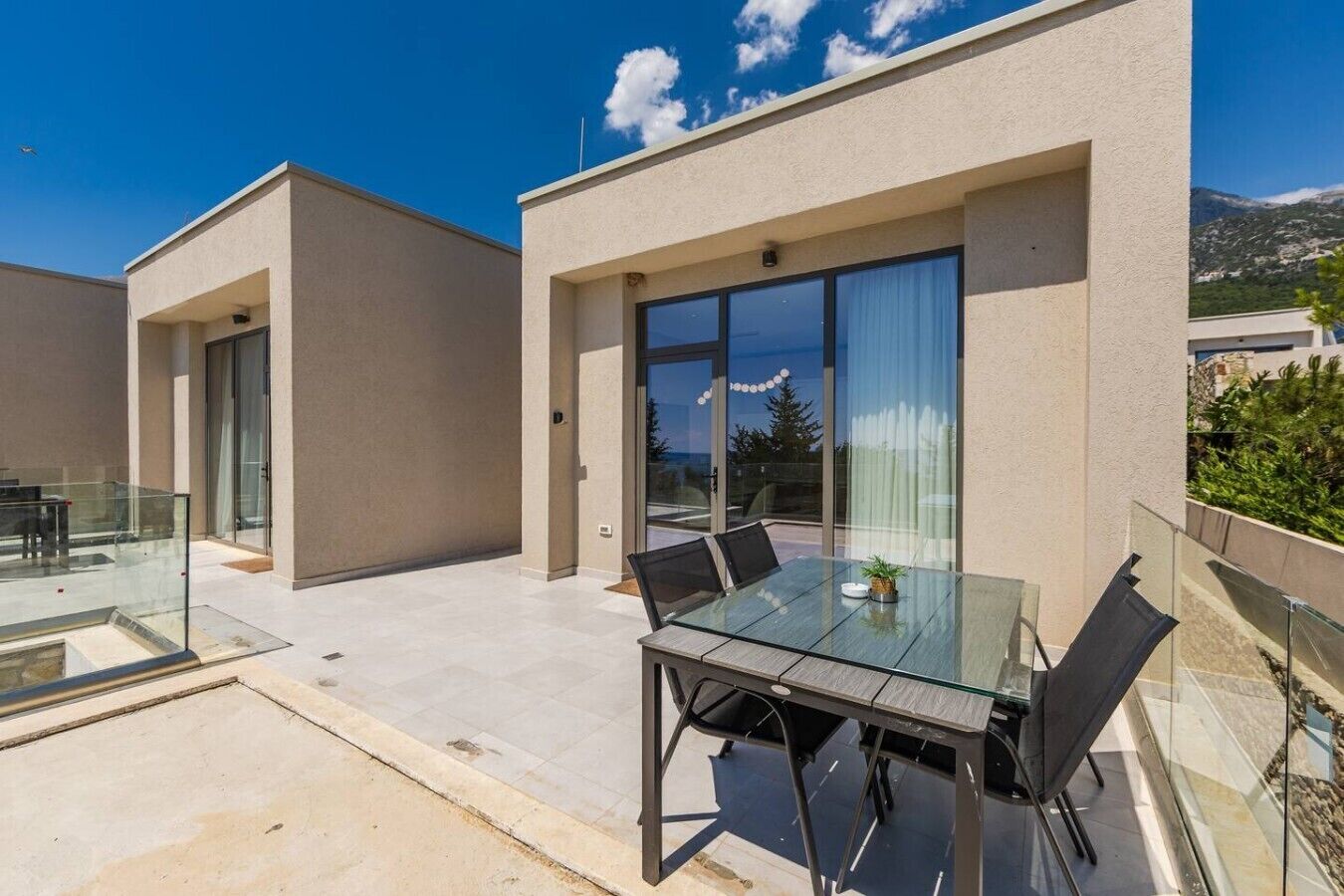 Un patio moderno al aire libre con una mesa y sillas de vidrio, balaustrada de cristal y vistas del paisaje circundante.