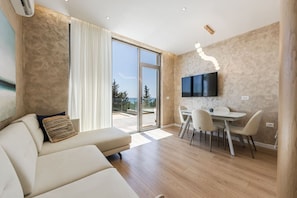 Ein modernes Wohnzimmer mit Sofa, einem Essbereich mit Tisch und Stühlen, einem an der Wand befestigten Flachbildfernseher und einer großen Schiebeglastür, die zu einem Außenbereich führt.