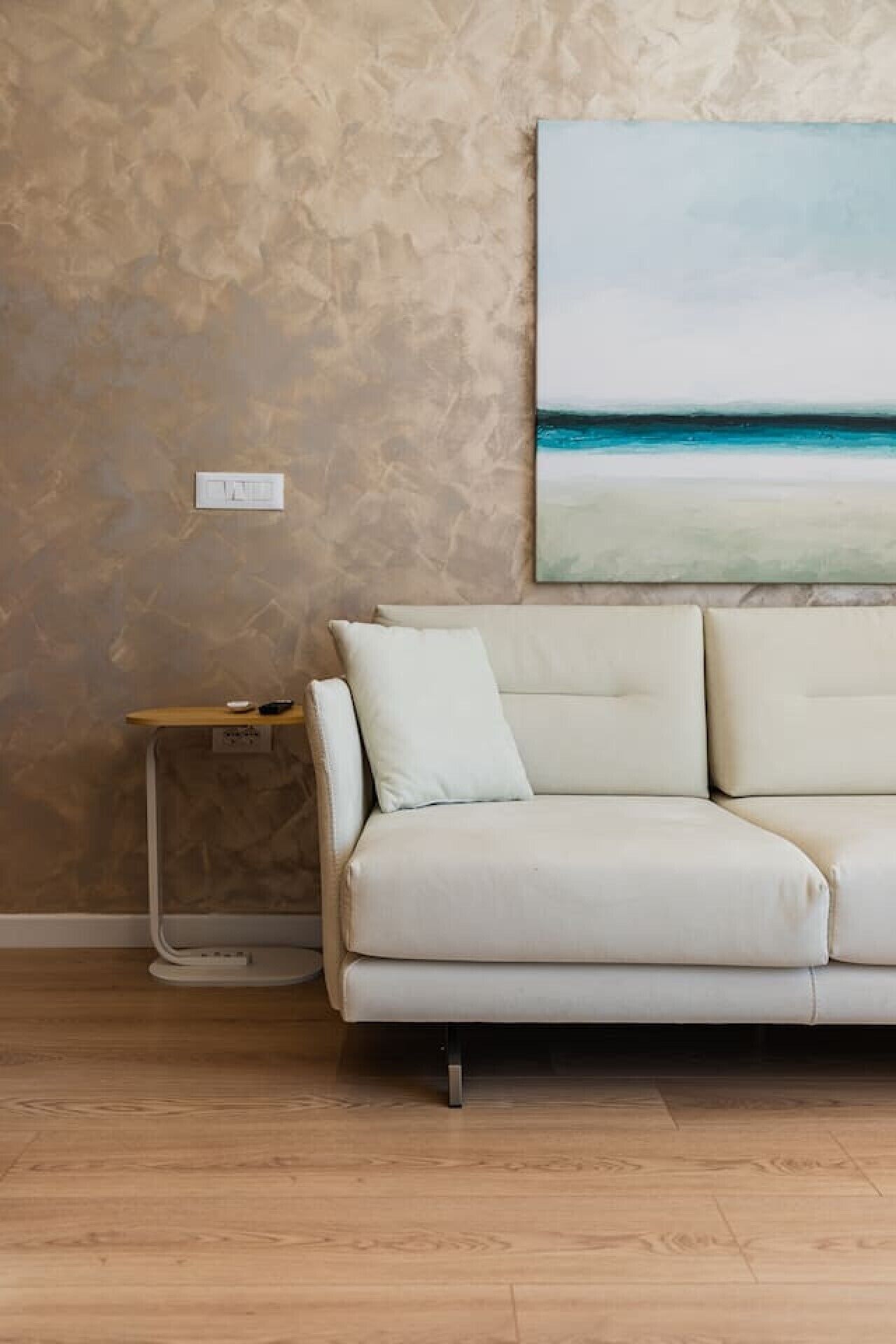 A beige sofa, a small wooden side table, and a painting of a beach scene on a textured wall.