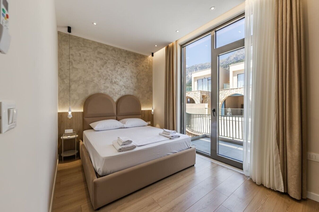 Una habitación de hotel con cama, mesita de noche, lámpara, un amplio ventanal corredero a un balcón y vistas de edificios.